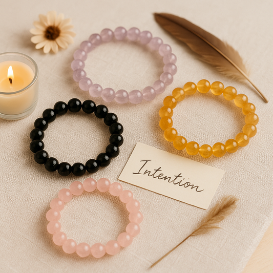Bracelets en pierres naturelles posés sur un tissu beige, représentant différentes intentions (amour, protection, abondance, sommeil), accompagnés d’une bougie, d’une plume et d’une carte portant l’inscription « Intention »
